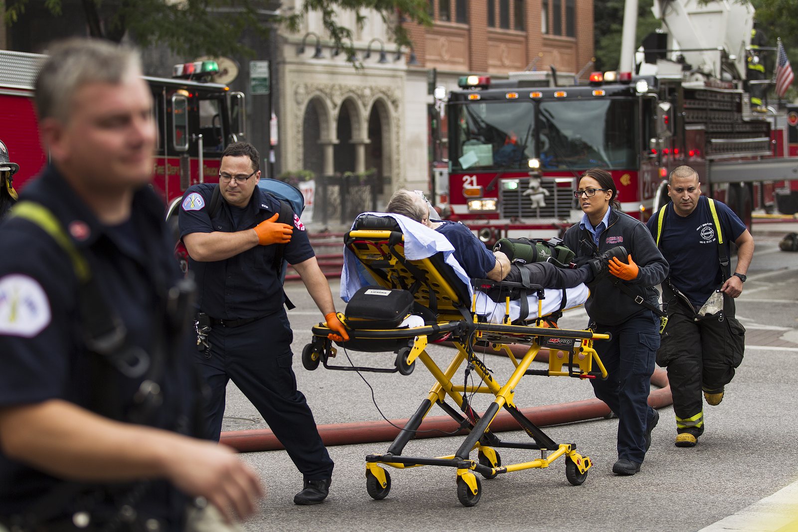 An EMS crew moves a patient on a stretcher past fire apparatus on a city street