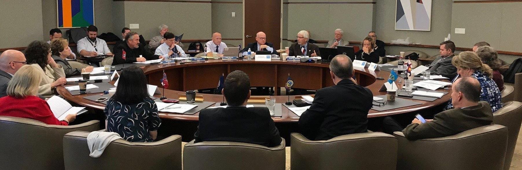 Commissioners seated around a conference table with state nameplates and flags during the first meeting of the Interstate Commission for EMS Personnel Practice.