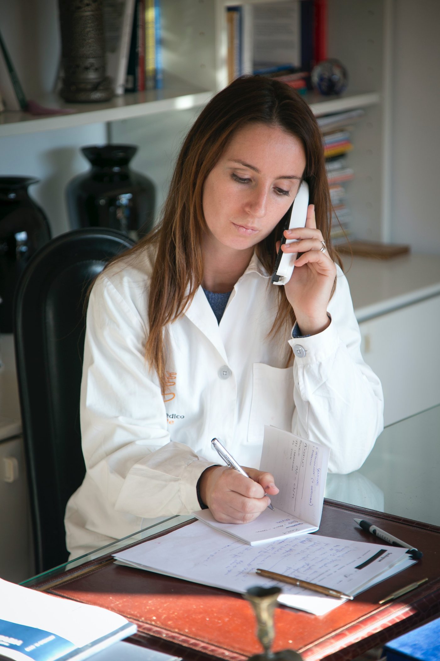 A physician in a white coat reviews notes while speaking on the phone at a desk