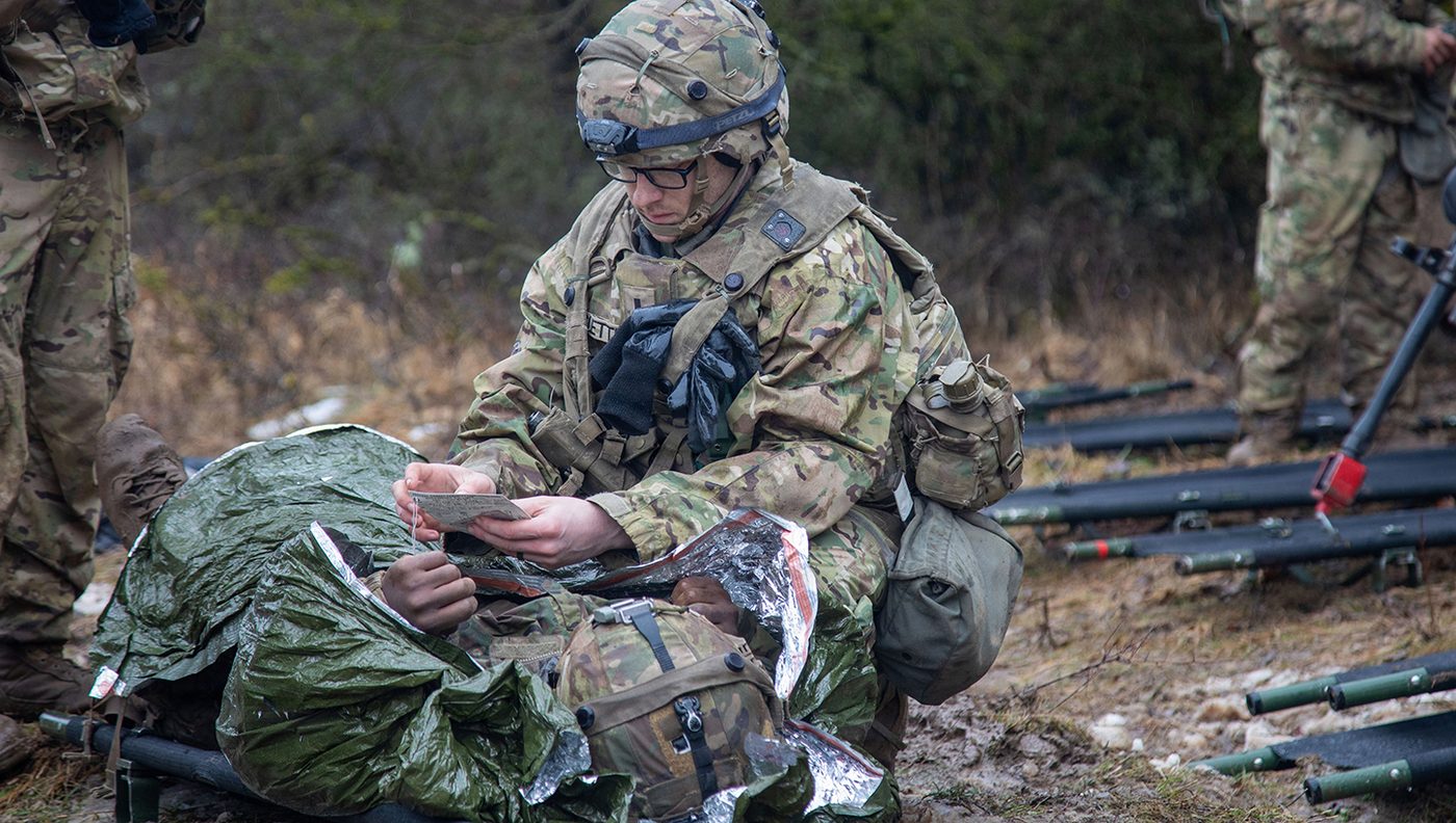 A soldier in camouflage provides medical care to a patient on a litter in the field